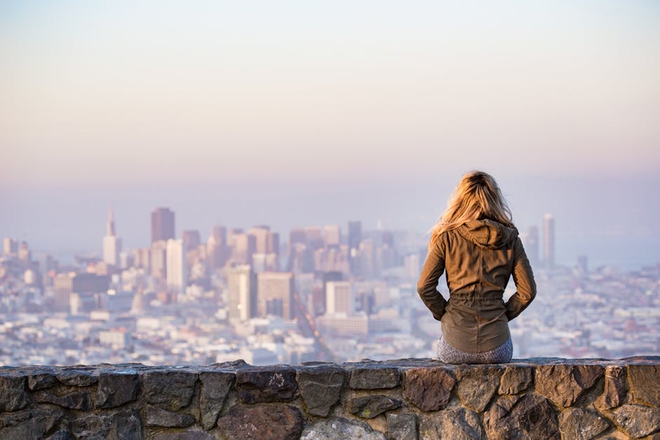 Travelling & Its Effect On Your Looks Woman sitting on rock wall looking out over cityscape 
