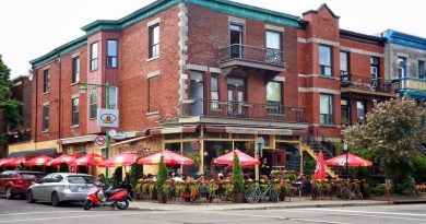 3 story red brick building with lovely landscaping outside, with tables and chairs and red shade umbrellas.