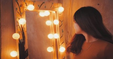 Woman with Brunette Hair and Wearing a brown top, looking into a lighted mirror -How To Like Yourself