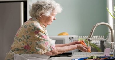 Elderly woman in floral dress and apron washing vegetables in kitchen sink - Aging in Place