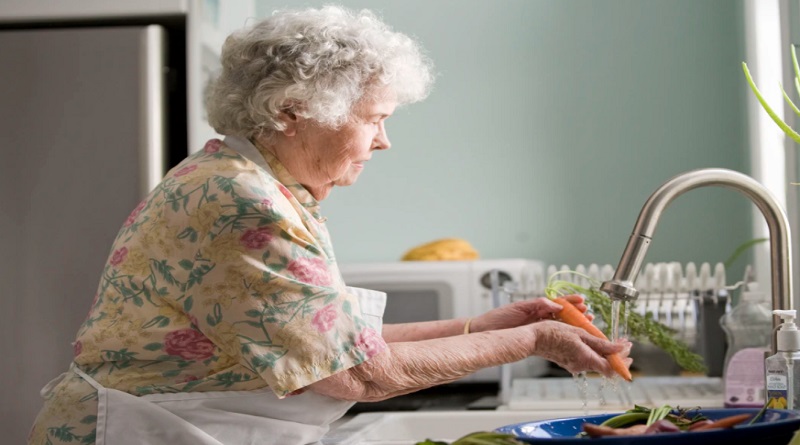 Elderly woman in floral dress and apron washing vegetables in kitchen sink - Aging in Place