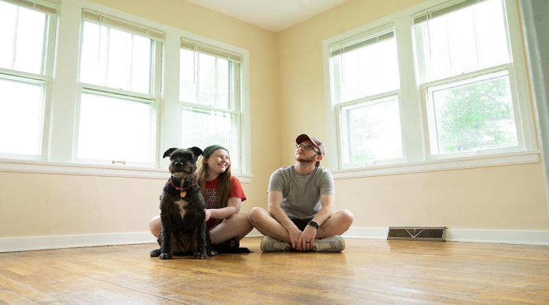 Up Sticks And Move Young couple and their dog sitting on the floor in a room with no furniture or drapes on the windows