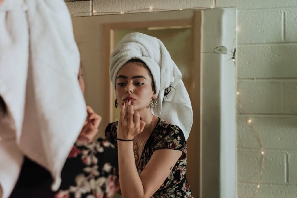 Personal Grooming Hacks Women with her hair up in a towel, looking into a mirror and applying product to her lips