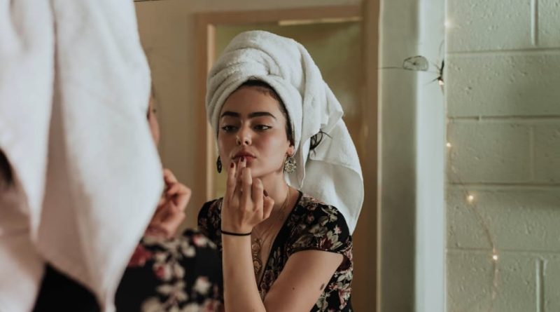 Personal Grooming Hacks Women with her hair up in a towel, looking into a mirror and applying product to her lips