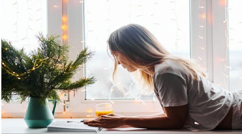 woman reading a book in front of window with fairy lights next to a vase filld with fir tree trimmings