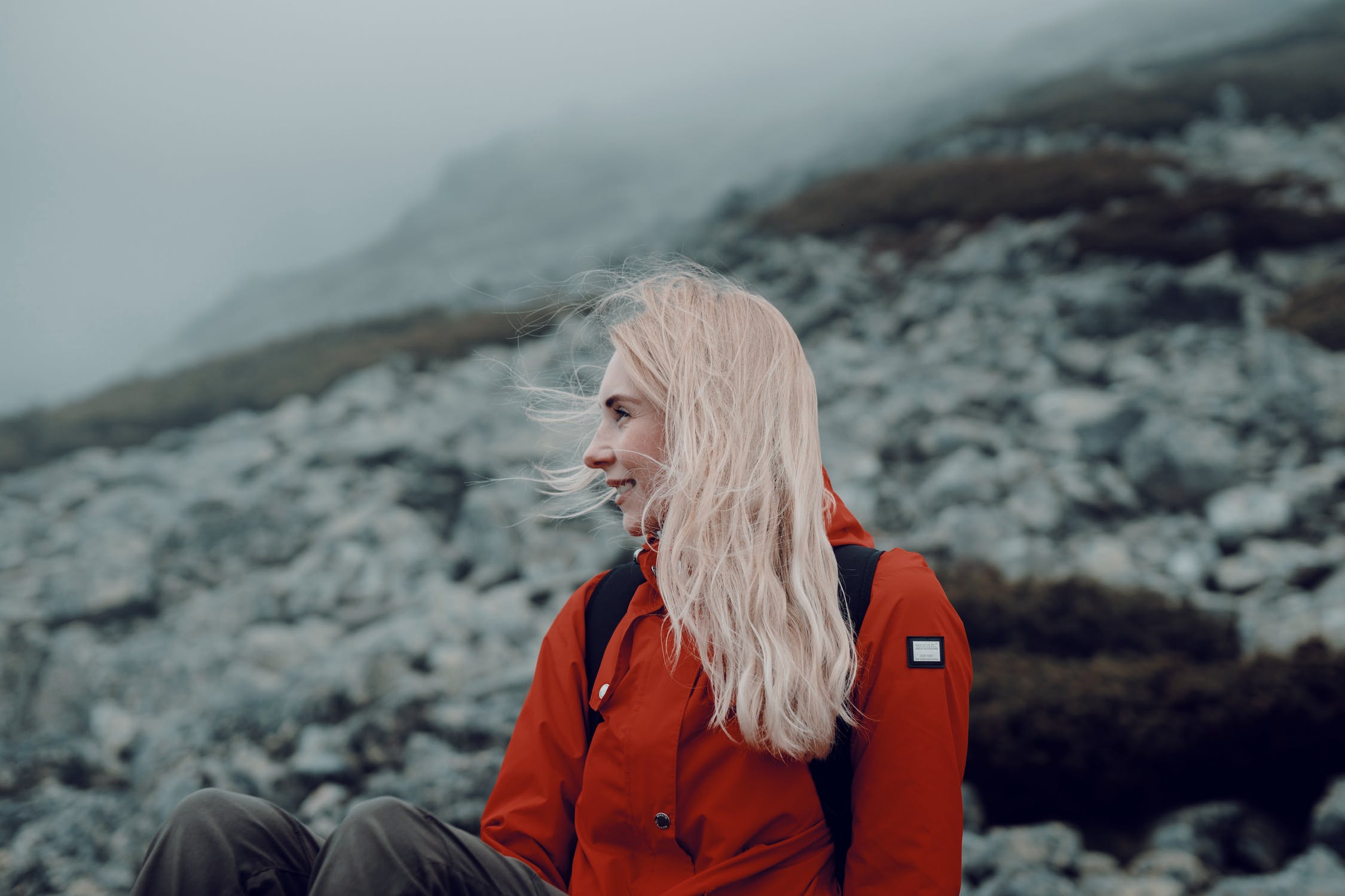 Woman with blonde hair wearing a red jacket sitting on a rocky hillside