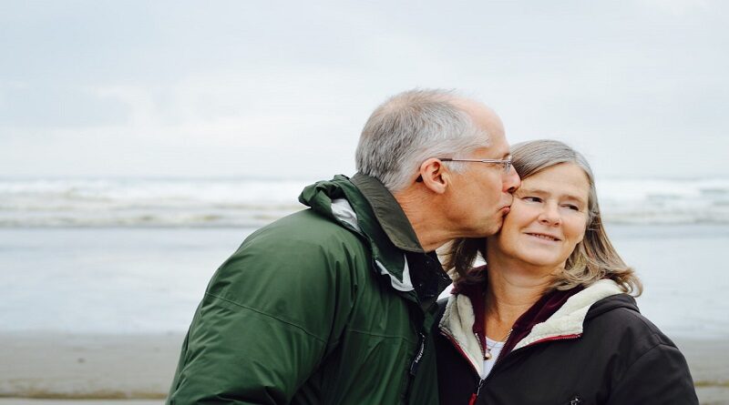 Ways You Can Give Back To Your Parents / Older couple on beach with man kissing the woman on the cheek