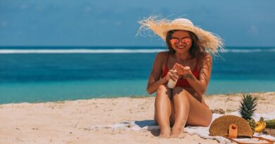 Looking After Your Skin in Summer / Woman in red bathing suit wearing a large straw hat stting on the beach putting on sunscreen