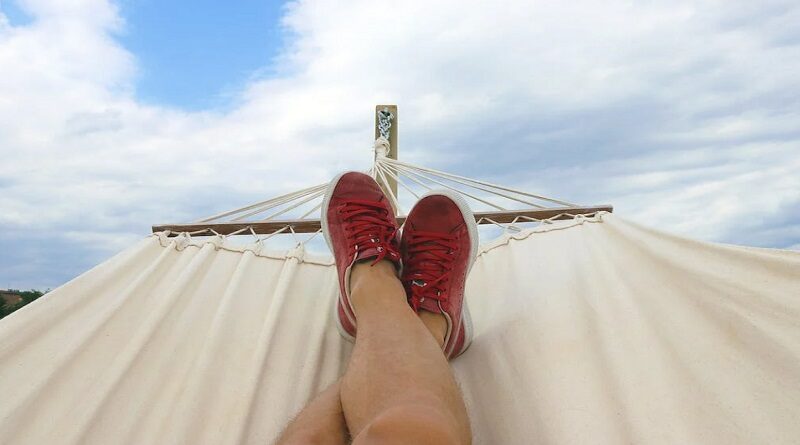 Mans legs and feet in red sneakers laying in a hammock under a blue and cloud filled sky