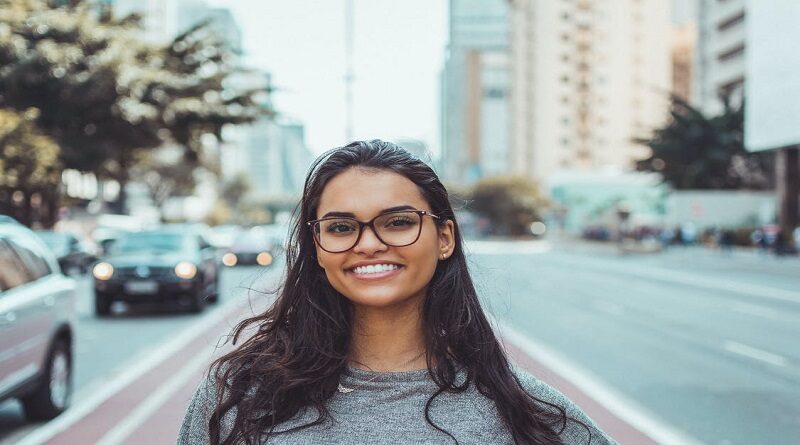 Dark haired woman with glasses smiling while standing in a center median of a busy street