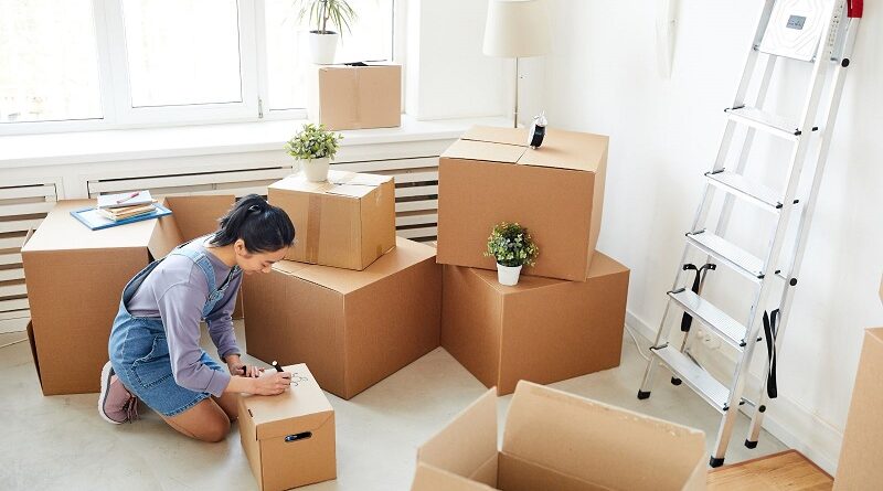 Packing for a Long Distance Move: Essential Items to Bring and Items to Leave Behind / Woman writing on a box surrounded by packed boxes for a move
