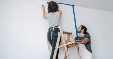 Man holding a ladder that a woman is standing on to tape off a wall for painting / Your Guide to Safely Painting Your Home
