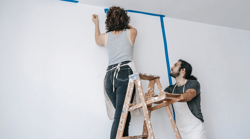 Man holding a ladder that a woman is standing on to tape off a wall for painting / Your Guide to Safely Painting Your Home