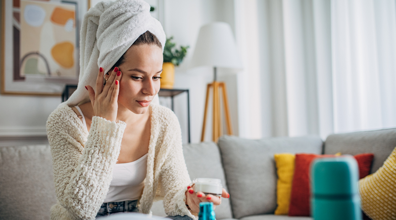 Woman with her hair up in a towel, sitting on a sofa putting cream on her face. / How Multi-Purpose Products Can Help Streamline Your Beauty Routine