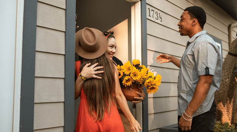 Couple being greeted by a woman at the door holding a pot of sunflowers / Guests Staying For The Summer? How To Prepare Your Home