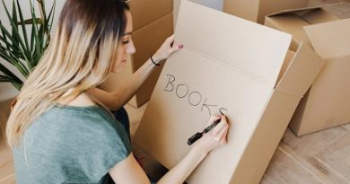 Woman sitting on the floor surrounded by boxes, writing the word books on one of them / Is Now A Good Time To Have A Clear Out?